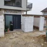 Unfertiger Eingang mit Betonblöcken und einer schwarzen Tür, überall verstreute Baumaterialien.