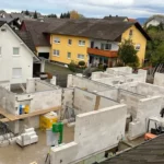 Teilweise gebautes Haus mit weißen Ziegelwänden, Baumaterialien und Häusern im Hintergrund.