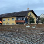 Drei Arbeiter stehen unter bewölktem Himmel auf einem Bewehrungsgitter auf einer Baustelle in der Nähe von Wohnhäusern.