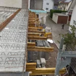 Blick auf eine Baustelle mit Stahlbewehrung, Holzbalken und Baumaterialien in der Nähe von Wohnhäusern.