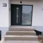 Moderne Haustür mit Glaseinsatz, vier Betonstufen und Hausnummer 34 auf einer weißen Wand.