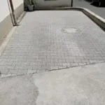 Kleiner gepflasterter Parkplatz mit einem runden Schacht, begrenzt durch niedrige Betonmauern und einer Topfpflanze in der Ecke.